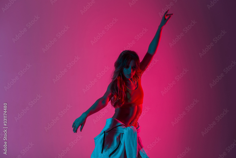 Sticker Sexy model sensual modern young woman in a T-shirt with a hoodie on the waist raises hands up in the studio with red color. Modern girl dancer posing and dancing in with room with amazing pink light.