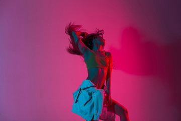 Fashionable young woman model in stylish youth clothes dancing in the studio with amazing bright neon pink color in disco style. Modern slim sexy girl dancer posing indoors with multi-colored light.