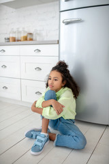 Dark-haired girl in jeans sitting near the fridge feeling hungry