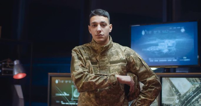 Portrait shot of Caucasian handsome young male soldier in camouflage uniform taking off cap and looking at camera in monitoring room. Man militarian posing in controlling office.