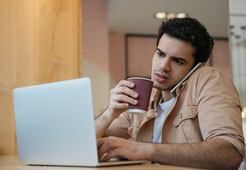 Portrait of pensive asian businessman doing multitasking, using laptop, answering calls, communication, drinking coffee in modern office. Indian man talking on mobile phone, working from home