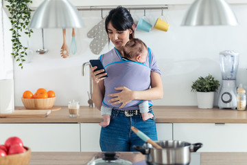 Pretty young mother with little baby in sling using her mobile phone in the kitchen at home. - Powered by Adobe