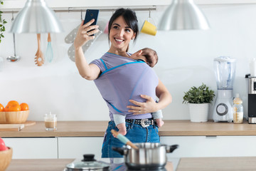 Pretty young mother with little baby in sling taking a selfie in the kitchen at home.