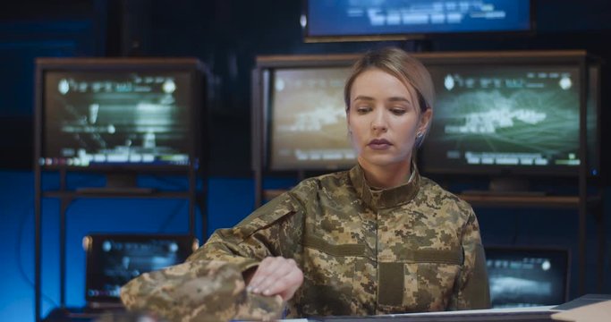Portrait of pretty Caucasian female soldier sitting at desk in monitoring room, taking on cap and looking at camera. Attractive woman in militarian uniform working in troops. Girl in army concept.