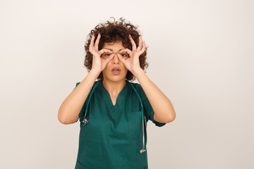 Playful excited medical female with dark long hair showing Ok gestures with both hands, pretending to wear spectacles, astonished to see something amazing. Emotions, body language and gestures.