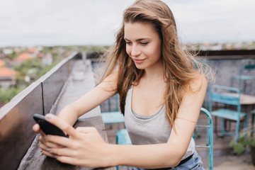 Interested white girl using smartphone on street background. Outdoor shot of carefree long-haired...