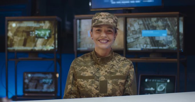 Portrait of beautiful Caucasian female soldier sitting at table in controlling room and smiling cheerfully to camera. Pretty woman in camouflage uniform and cap working in troops. Girl in army concept