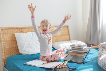 Happy Caucasian girl child sitting in bed learning online on Internet. Virtual class lesson on video at self isolation in home. Distant remote video education. A modern school study for kids.