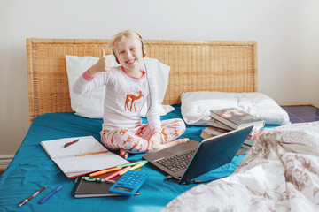 Fototapeta na wymiar Happy Caucasian girl child sitting in bed learning online on Internet. Virtual class lesson on video at self isolation in home. Distant remote video education. A modern school study for kids.