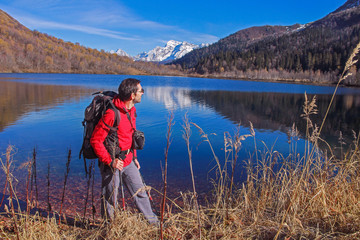 Fototapeta na wymiar A middle-aged man in a red jacket and glasses with a backpack for tourism on the background of the lake which reflects the snow-capped mountains.