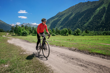 Fototapeta na wymiar A young smiling girl on a cyclocross bike rides along a winding mountain road against a background of green forest and mountains with glaciers and snow on the tops