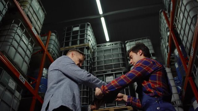 From below tracking shot of businessman and material handler signing contract and shaking hands in industrial storage