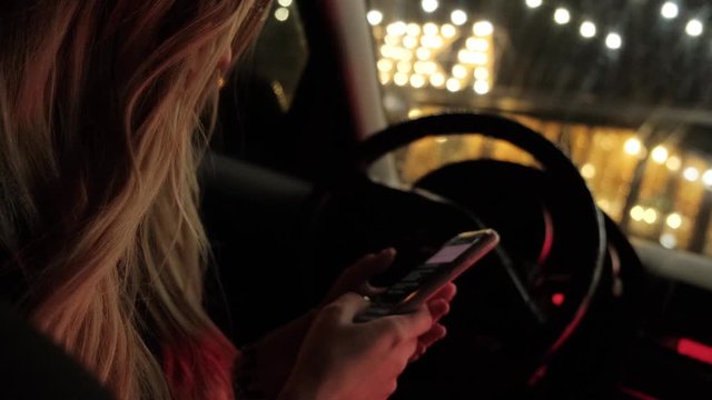 Blonde in a car with a smartphone in her hands. Girl is typing closeup. Night city. Slow motion.