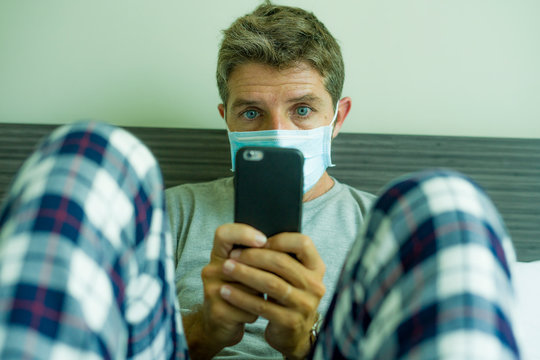 Virus Outbreak - Dramatic Portrait Of Sick Man In Medical Facial Mask Sitting On Bed Scared And Worried Infected By Coronavirus Suffering Lockdown Alone At Home In Quarantine