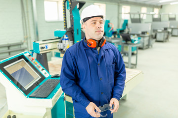 CNC machine operator in the Hard  white Hat Walks Through Light Modern Factory While Holding tablet. Successful, Handsome Man in Modern Industrial Environment.