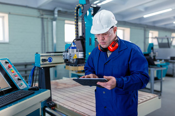 CNC machine operator in the Hard  white Hat Walks Through Light Modern Factory While Holding tablet. Successful, Handsome Man in Modern Industrial Environment.