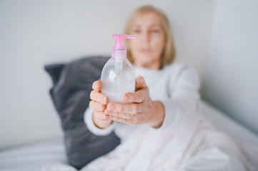 Hand sanitizer bottle senior woman using dispenser gel rub for corona virus hands hygiene coronavirus COVID-19 pandemic prevention. Patient lying in bed at home quarantine