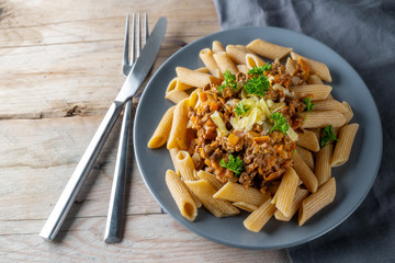 wholemeal penne noodles with a sauce of minced meat and carrots, parmesan cheese and parsley garnish, cutlery on a rustic wooden table, copy space