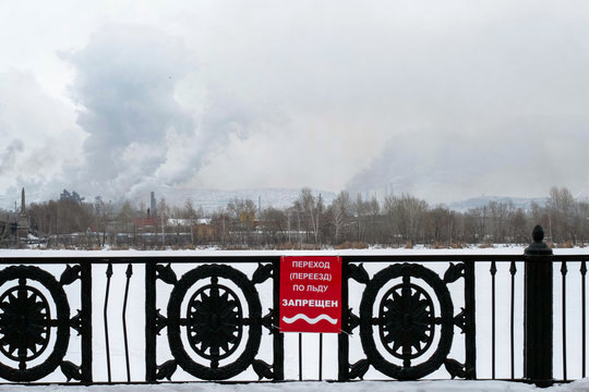 The announcement on the fence by the river. The message is fixed on the fence. Words: crossing (moving) on ice is prohibited. View of the river, factory, chimneys and smoke