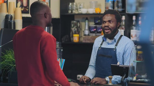 black bearded man in apron stands behind bar counter and talks with client from native country and doing high-five against bar