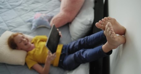 Little girl watching movie in bedroom. Barefoot young people lying in bed at home. Happy redhead female child looking at video with laptop computer and smiling - Powered by Adobe