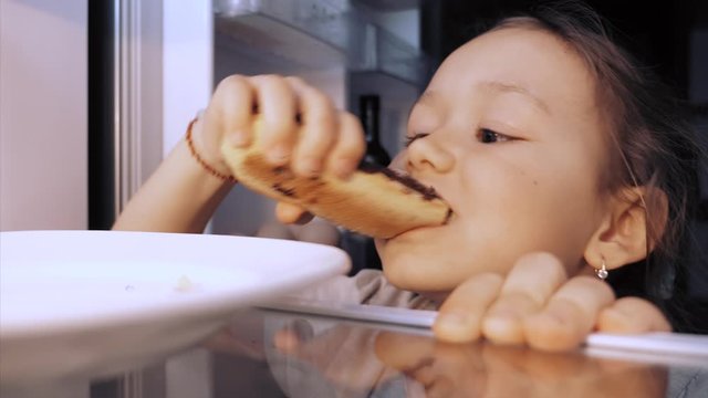 Little child girl is opening a fridge at night, taking an eclair and eating it furtively. View from refrigerator.