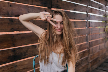 Fototapeta premium Amazing girl with brown hair posing with peace sign in good spring day. Outdoor shot of glad caucasian female model laughing on wooden background.