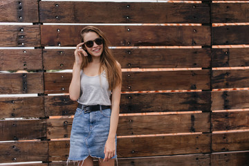 Inspired caucasian girl in trendy clothes posing near wooden wall. Photo of spectacular lady in stylish denim skirt smiling to camera.