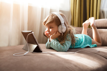 Child girl in headphones is using a digital tablet while lying on the sofa at home. Training...