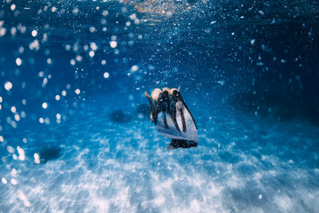 Woman freediver glides with fins over sandy sea bottom and bubbles.