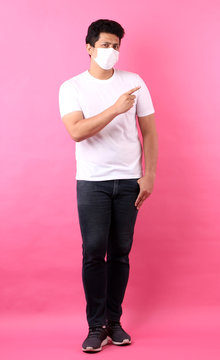 Full Body Portrait Of Handsome Asian Man Wearing A Mask Is Sick Pointing Finger Isolated On Pink  Background In Studio.