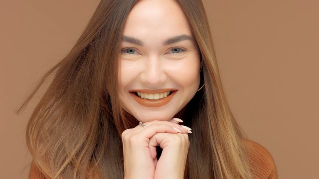 closeup portrait of smiling young influencer watching to the camera on brown background with har blowing