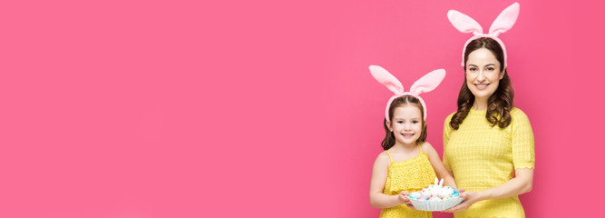 panoramic shot of happy mother and daughter in bunny ears holding easter eggs isolated on pink