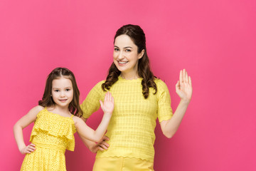 happy mother and daughter waving hands isolated on pink