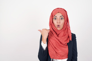 Horizontal shot of pretty young joyful shocked female points with thumb away, indicates something on blank wall isolated over gray background. Check this out. Ethnicity concept.