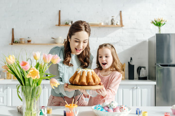 excited mother and child looking at easter bread near easter eggs, decorative rabbits and tulips
