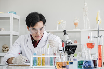 A male scientist with black hair wearing white coat and protective glassware holding up test tube in a laboratory setting with solutions and microscope.