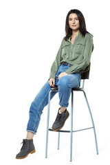A young woman in jeans, boots and a khaki shirt is sitting on a high chair. Isolated on white.