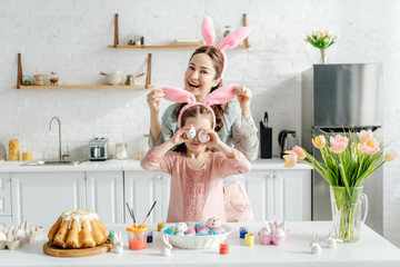 happy mother touching bunny ears on daughter covering eyes with chicken eggs near easter bread and tulips