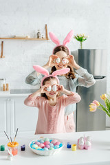 Happy mother and daughter in bunny ears covering eyes with easter eggs near tulips