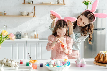 selective focus of happy mother touching bunny ears on daughter painting chicken egg  near easter bread