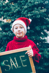 A teenager in a Santa Claus costume holds a gift. Christmas tree for the New year. Child, boy smiling. It's Snowing, snowflakes. Advertising, discounts, sales.