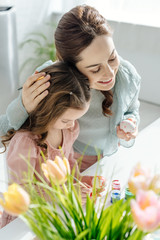 Obraz na płótnie Canvas selective focus of happy mother hugging cute daughter painting easter egg near tulips