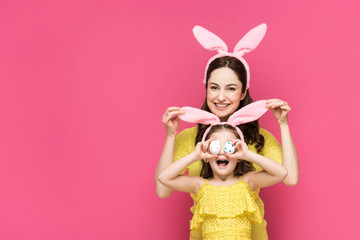 happy mother touching bunny ears of daughter covering eyes with easter eggs isolated on pink