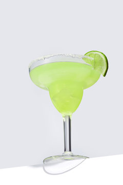 Margarita cocktail with ice cubes is contained in a margarita glass with a lime slice and salt on the rim and isolated on the table edge. The showy illustrative picture is made on the gray background.