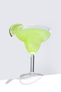Margarita cocktail with ice cubes is contained in a margarita glass with a lime slice and salt on the rim and isolated on the table edge. The showy illustrative picture is made on the gray background.
