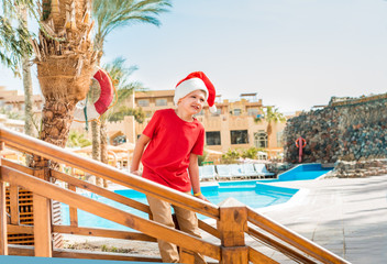 Santa claus on a vacation on a tropical beach.Palm trees and ocean, sea vacation.Cute little boy in santa costume. Winter trip. Summer hot sun, blue sky and white sand.Hotel in Egypt, Turkey or UAE