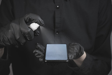 Cleaning the phone with a sanitizer, total black concept. Hands in black gloves spray sanitizer on the smartphone screen. Virus spread prevention concept