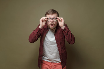 Surprised bearded man, in a burgundy shirt, in amazement holds glasses in hands and looks with big...