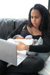 A young beautiful African American mom works remotely from home for work with her laptop on her lap and baby infant girl in her arms as she sits on a black leather sofa by a window.